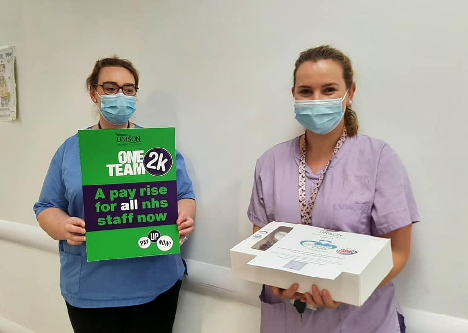 Two nurses, one holding a placard that says 'a pay rise for all NHS staff now' and one carrying a box of cakes