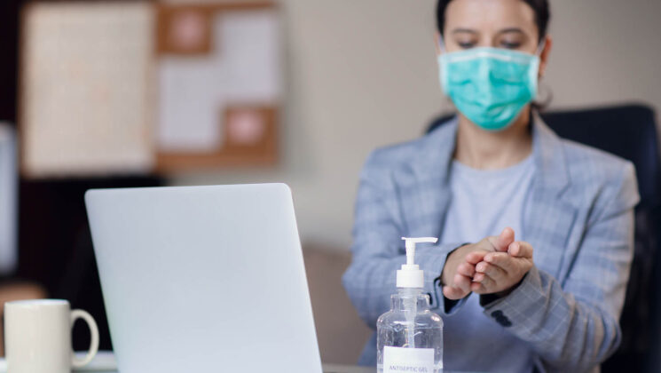 woman with mask on in front of laptop, sanitising her hands