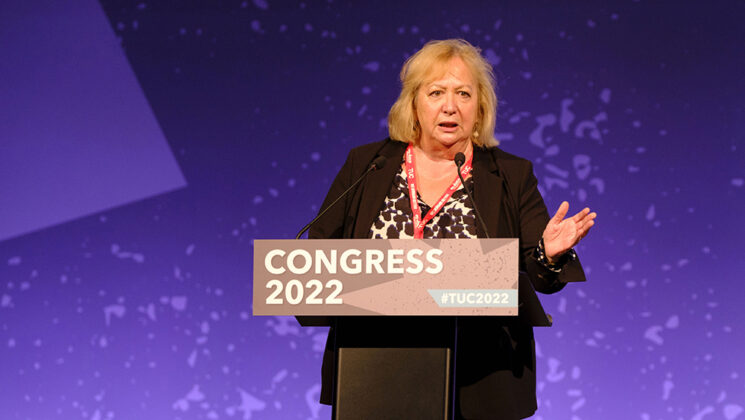 Christina McAnea addressing TUC congress in Brighton. She is wearing a black jacket over and black and white top and is pictures at the congress rostrum, her left had raise, and against a dappled purple background