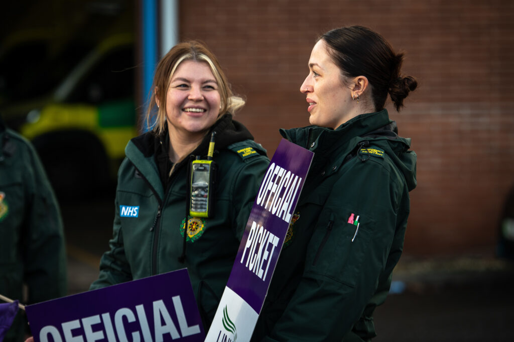 Two paramedics smile and hold 'official picket' placards outside Longley ambulance station, Sheffield