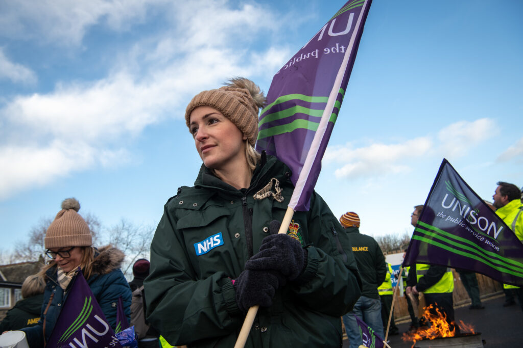 A woman ambulane worker holds a flag over her should with a small brazier fire and blue sky in the background