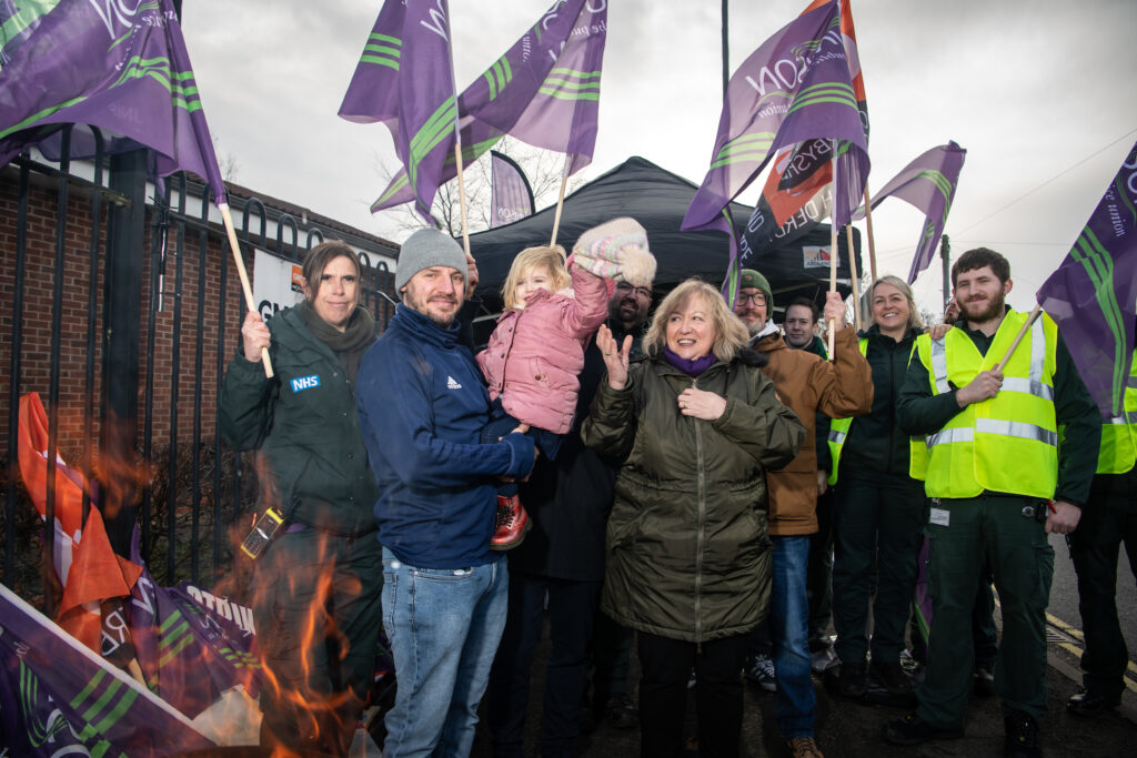 Christina stands on York ambulance station picket line behind a flaming brazier with strikers holding, including one holding a young child in their arms