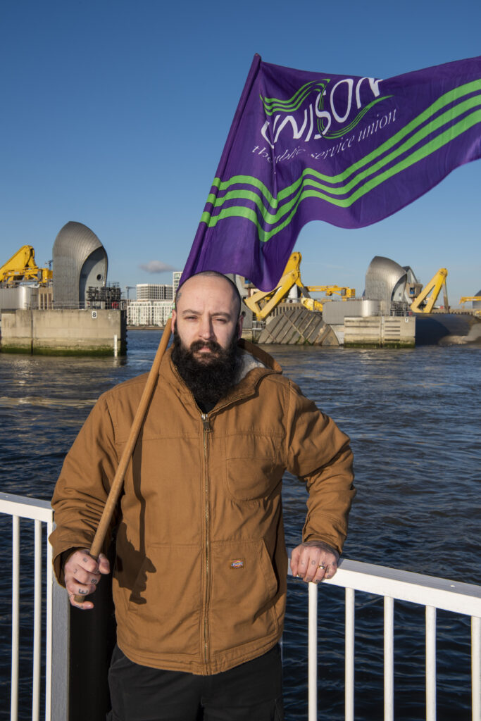 A striking UNISON environment agency member standing in front of the Thames Barrier with a UNISON flag