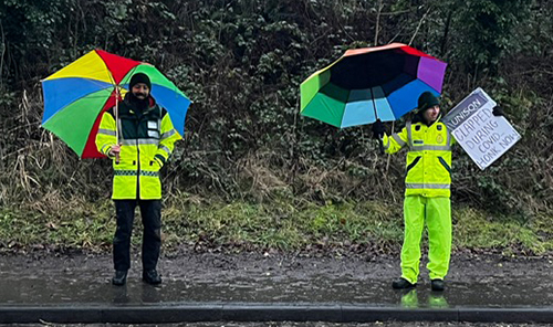 Two North West Ambulance Service pickets with bright umbrellas and a placard, by a road