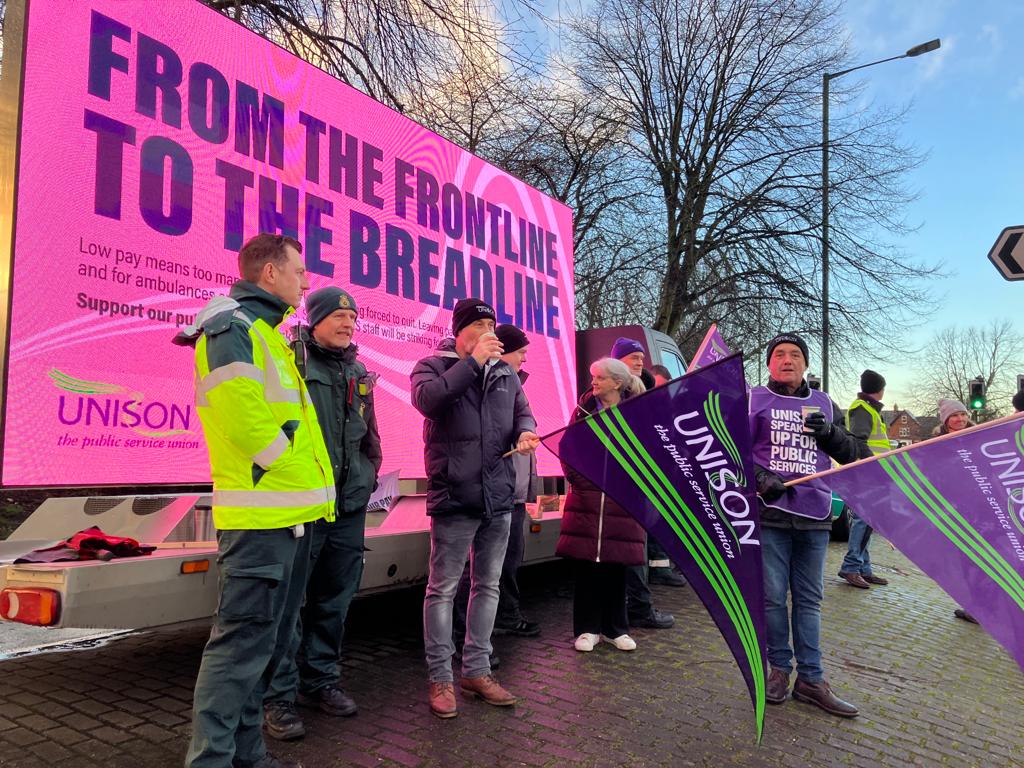 Ambulance strikers at Chester le street stand in front of a pink ad van which says "from the frontline to the breadline"