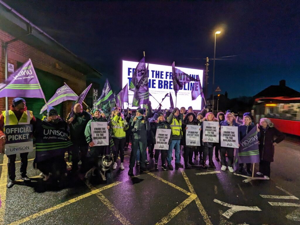 Christina McAnea with strikerson the ambulance picket line in Gateshead after dark in front of an ad van reading "from the frontline to the breadline" 