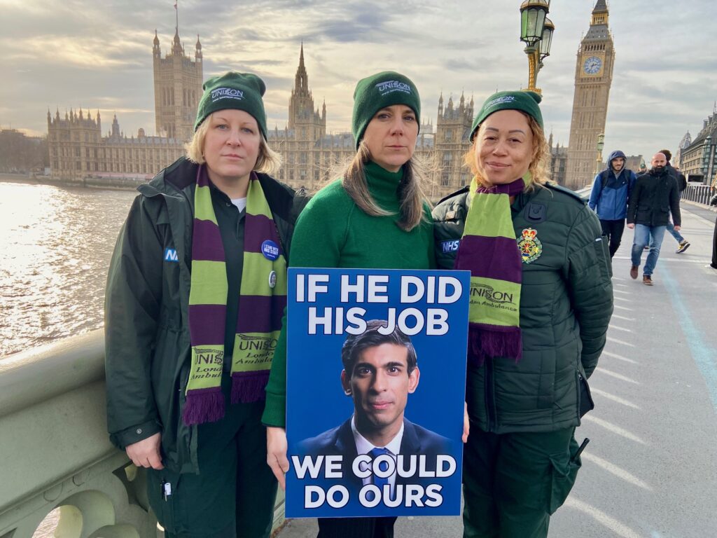 Sara Gorton stands on Westminster bridge with two London Ambulance Service strikers - with Parliament in the background, holding a placard with a picture of Rishi Sunak, saying: "If he did his job, we could do ours."
