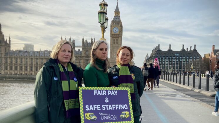 Sara Gorton stands on Westminster bridge with two London Ambulance Service strikers - with Parliament in the background, holding a placard saying:"Fair pay & Safe Staffing"