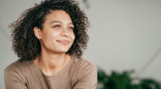 Young woman with curly hair wearing a light brown long sleeved top smiles calmly and looks into the distance