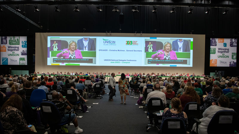 Photograph from back of national delegate conference hall, showing members in seats and the backdrop showing image of Christina McAnea speaking at the podium