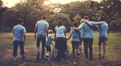 7 people, all in the same blue shirts walk away from camera across a field, the sun setting in the distance. It is a diverse group with a child and a person in a wheelchair. Some of them have their arms around each others shoulders